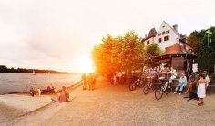 Der Weinprobierstand befindet sich direkt am Eltviller Rheinufer. Personen sitzen am Weinstand, andere sitzen direkt am Rheinufer oder gehen dort spazieren. Im Hintergrund geht die Sonne hinter den Platanenbäumen unter.